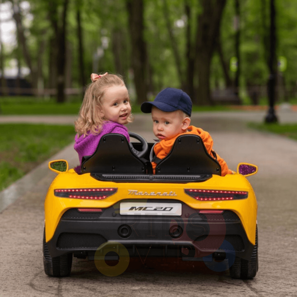 Two young children are seated in a red XXL Maserati MC20 ride-on car on a paved park path. The girl, with curly hair, wears a purple jacket; the boy, in an orange outfit, dons a blue cap. Green trees and a lamppost add to the serene background.