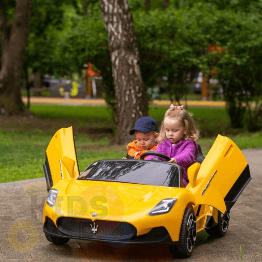 Two young children enjoy the Experience Luxury: Yellow XXL 24V Maserati MC20, a premium 2-seater with RC, leather seats, and rubber tires. The left one wears an orange shirt and cap; the right one sports a pink jacket. They are parked on a path in the park, surrounded by trees and grass.