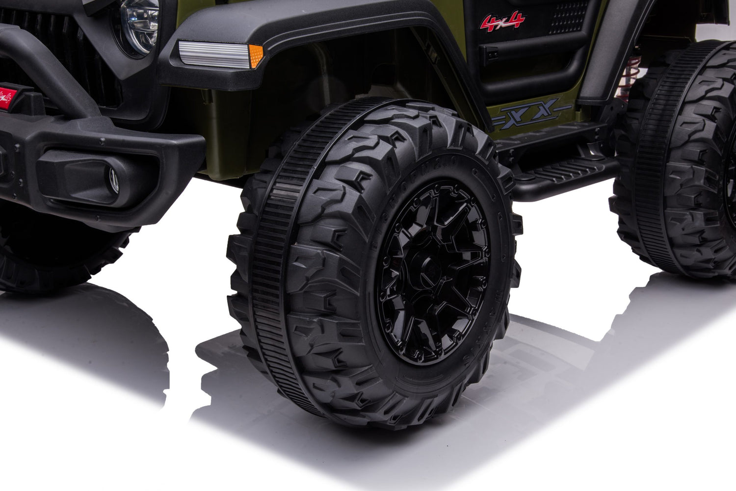 Close-up of a Black 24V/2WD Xtreme Edition Ride on Trucks rugged eva wheel with deep treads on a glossy white surface. Part of its dark green and black body, complete with visible decals and leather seats, is also shown.