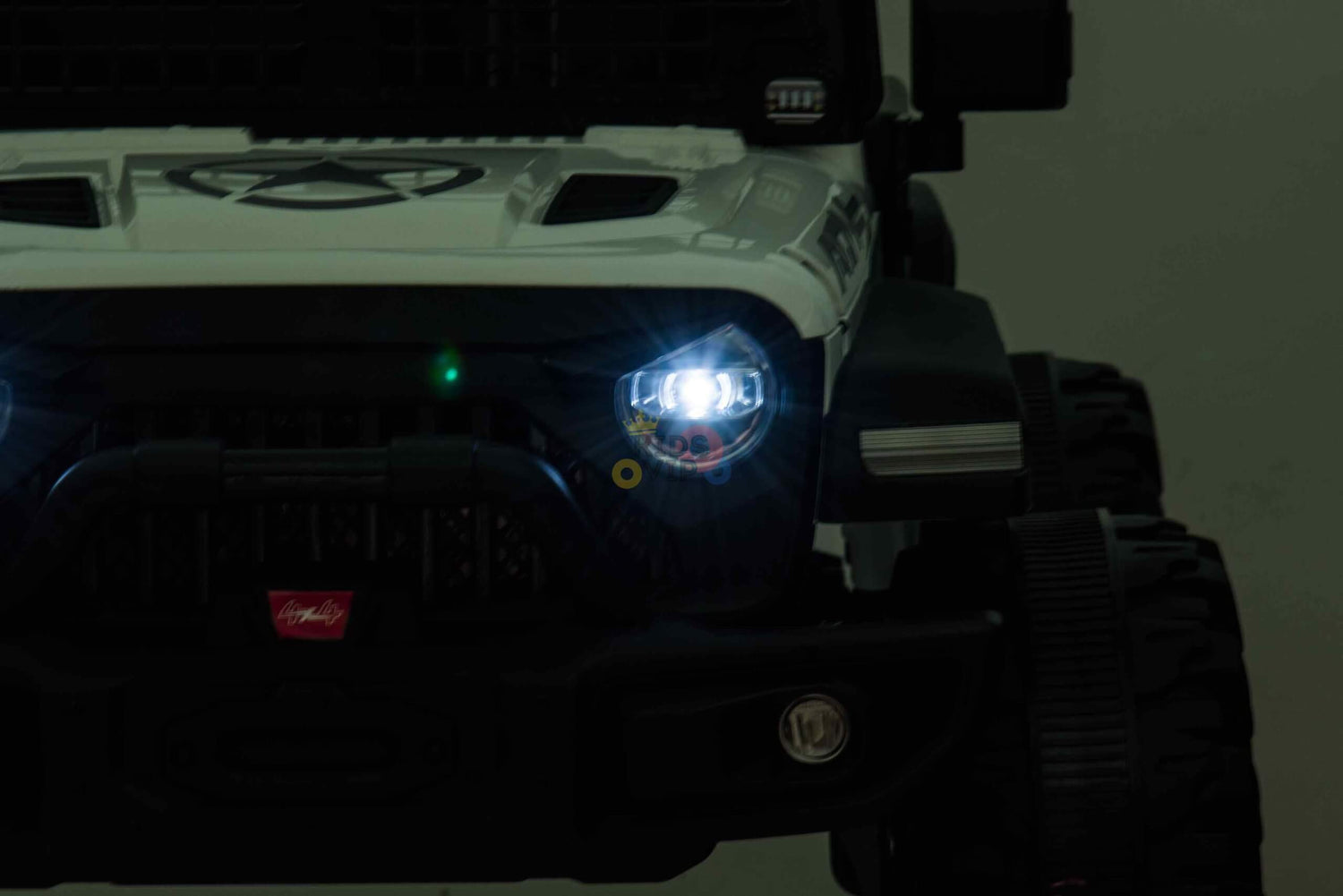 A close-up of the White 24V/4WD Xtreme Ride on Truck showcases its glowing headlights. The black and white toy jeep highlights rugged details like a grille and large Eva wheels, with bright lights contrasting against the dark background.