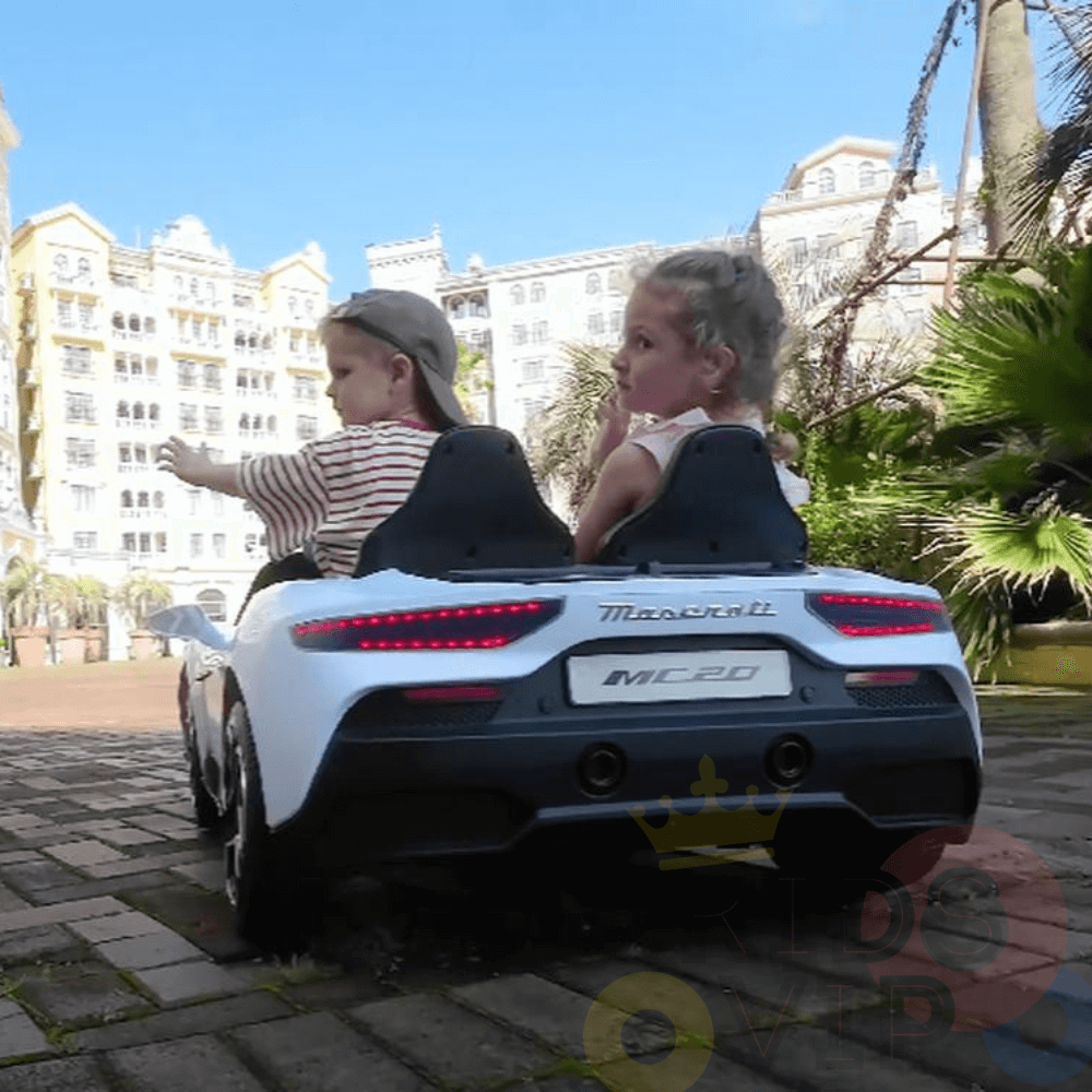 Two kids enjoy the Experience Luxury: Blue XXL 24V/105W Maserati MC20, a premium 2-seater with leather seats and rubber tires, driving it on a paved path near greenery and an elegant building.