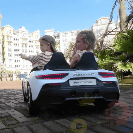 Two children have a luxury ride-on experience in the Experience Luxury: White XXL 24V /105W Maserati MC20 toy car, cruising on a brick path. One points forward while the other looks to the side, against a backdrop of a large building and palm trees on a sunny day.