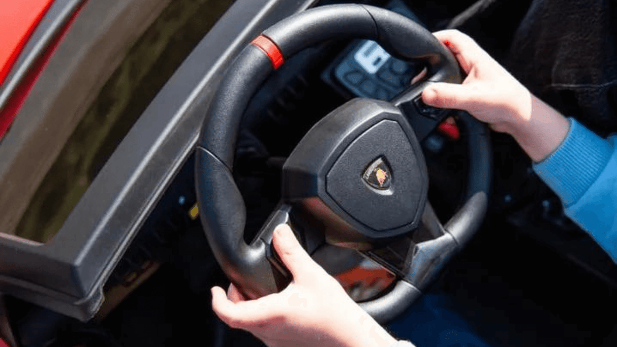 Hands grip the black steering wheel of a Blue Giant Official Limited Edition Lamborghini SV 24V/180W ride-on car, featuring a logo with a charging bull and a dashboard with controls and digital screen. Luxurious eco-leather seats are complemented by the person’s light blue sleeve.