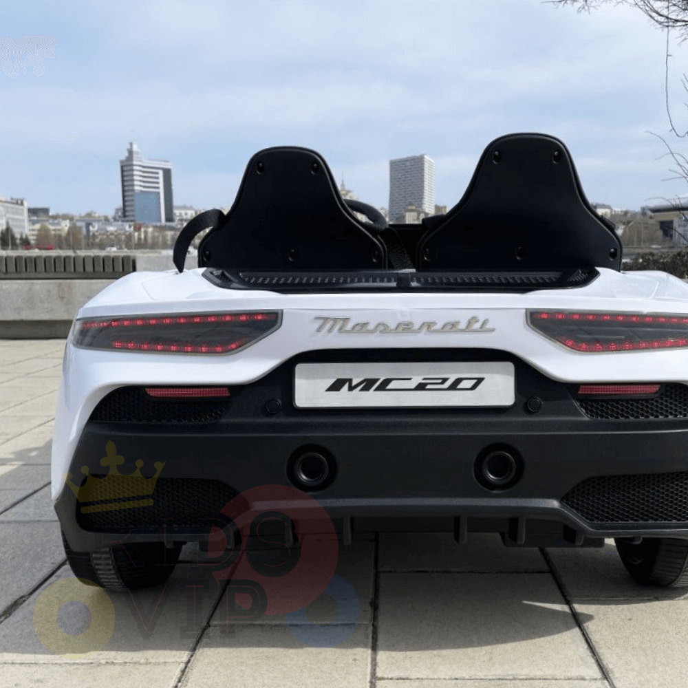 Rear view of a White XXL 24V Maserati MC20 ride-on car on a paved area, highlighting its unique taillights and dual exhausts. Against city buildings under clouded skies, this premium 2-seater with RC, leather seats & rubber tires epitomizes high-performance elegance.