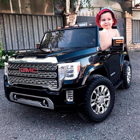 A small child in a red hat joyfully sits in a black 12V/2 Seater GMC Sierra ride-on car, gripping the high-quality steering wheel as the truck rests on gravel before a metal-sided building.