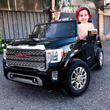 A smiling child is enjoying a shiny black 12V/2 Seater toy GMC Sierra on gravel, wearing a red bucket hat and no shirt, with a building and tree in the background.