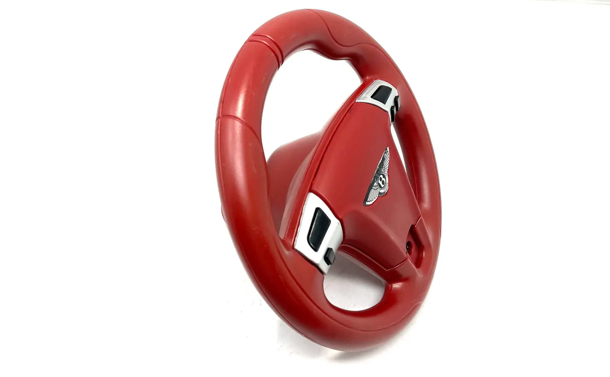 The 12V Bentley Sport GT Steering Wheel features a racing theme with red color, central emblem, and black and white accents on the grips. Perfect for young racers, its stylishly angled against a white background. Dont miss this final sale at KidsVipOnline!.