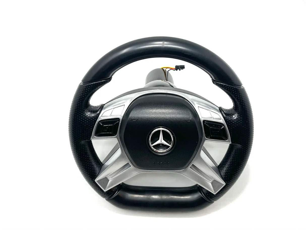 The 12V Unimog Steering Wheel, featuring a black and silver design with a metallic emblem at its center, is displayed on a white backdrop, highlighting the sleek aesthetic of KidsVIPOnlines Final Sale ride-on car collection.