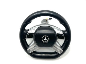The 12V Unimog Steering Wheel, featuring a black and silver design with a metallic emblem at its center, is displayed on a white backdrop, highlighting the sleek aesthetic of KidsVIPOnlines Final Sale ride-on car collection.