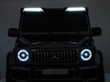 A Black Mercedes G63 XXL 24V Kids Ride-On Truck – Platinum Edition, featuring MP4, dual batteries, 4WD, EVA tires & leather seats, is parked in a dimly lit space. Bright LED headlights and a roof light bar illuminate its front grille with a circular emblem; plate reads DK-G63.