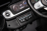 Close-up of a Black Mercedes G63 XXL 24V Kids Ride-On Truck dashboard, featuring a steering wheel, round air vent, digital display with gauges, and control panel with USB ports and radio display. The black interior with silver accents enhances the luxurious feel of this Platinum Edition ride-on.