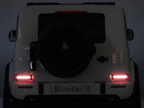 The rear view of a white Mercedes-Benz G-Class SUV, illuminated taillights echoing the Black Mercedes G63 XXL 24V Kids Ride-On Truck – Platinum Edition. A spare tire is mounted on the back door, with a G 63 badge near the right taillight. The license plate reads DK-G63.