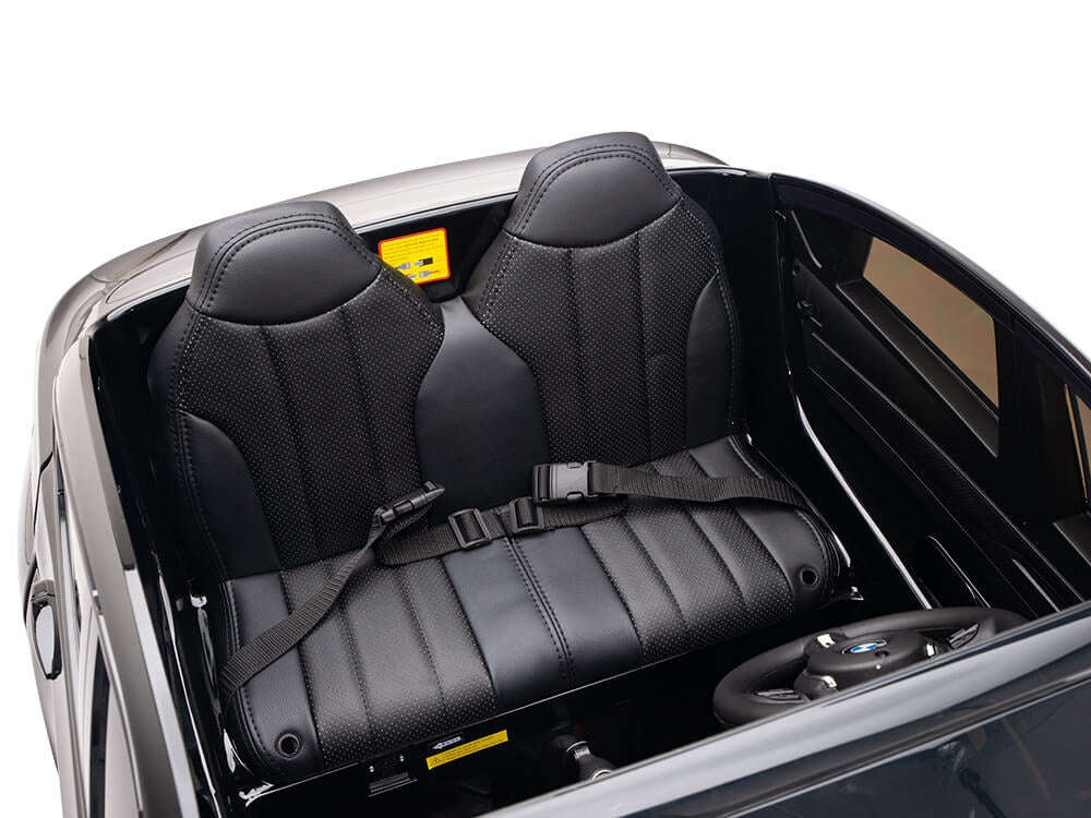 The image displays the interior of the Black 12V Licensed XXL BMW X6 Ride On Car with Remote Control, featuring two black leather-look seats with detailed stitching and seat belts. The dashboard includes a logo-adorned steering wheel and control panel, surrounded by stylish door frames.