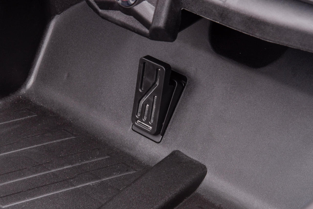 Close-up of a black pedal with a textured surface in the pink 2x24V/4WD Official Can-Am Maverick Ride on Buggy, LX Performance, mounted against a dark interior panel. The pedal has a slight rectangular pattern, surrounded by plastic trim against a grooved floor mat in this sleek vehicle.