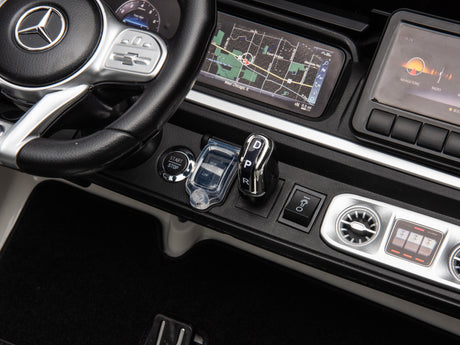 Close-up of a White 2x24V/4WD Platinum XXL Mercedes G63 Ride-On Truck dashboard featuring a logo-emblazoned steering wheel, infotainment screen with map, start/stop button, gear shift, and audio-climate control panel. Enjoy remote control access to this 4-wheel drive adventure.