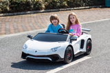 Two children sit in a White 24V/180W Lamborghini SV Limited XXL Edition on a paved road. The child on the right grips the wheel, both smiling amid greenery and more pavement, relishing their plush eco-leather seats at speeds up to 10MPH.