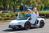 Two children are in a White 24V/180W Lamborghini SV Limited XXL Edition ride-on car with sleek lines and stylish open doors. Smiling and posed as if driving at up to 10MPH, they are outdoors amidst lush greenery with parked cars in the background.