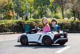 Two children zoom down a park path in their White 24V/180W Lamborghini SV Limited XXL Edition, iconic against lush greenery. The driver in pink and passenger in blue enjoy the thrill on eco-leather seats, adding luxury to their playful adventure at speeds up to 10MPH!.