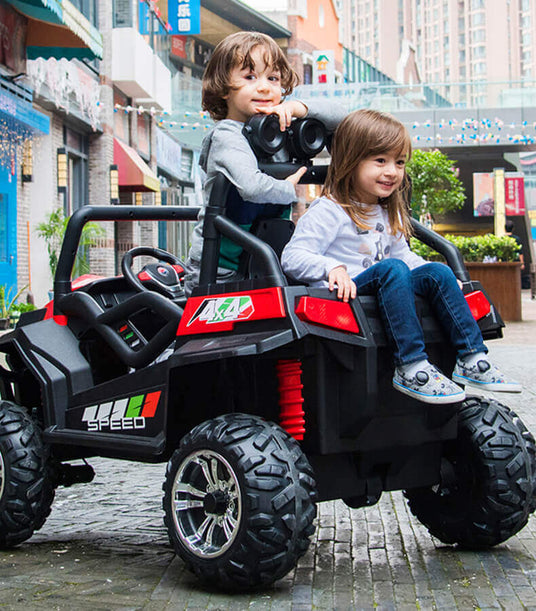 Two children sit in a toy car, one holding binoculars. They are on a cobblestone street in an urban setting, surrounded by buildings and greenery. The toy car is designed as a mini off-road vehicle.