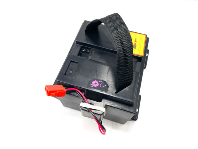 The 2x12v Complete Battery for the 12v/2 Seater GMC Sierra is black with a strap handle, a red connector plug, and a power symbol decal. A visible yellow warning label ensures its ready to power fun adventures.