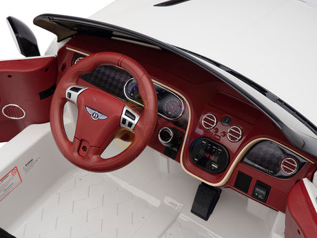 The White 12V Bentley GTC Kids Licensed Ride On Car features red and white interior detailing, a leather seat, a red steering wheel, dashboard gauges, various buttons, and a small door speaker. Its floor showcases a textured white pattern for an authentic ride experience.