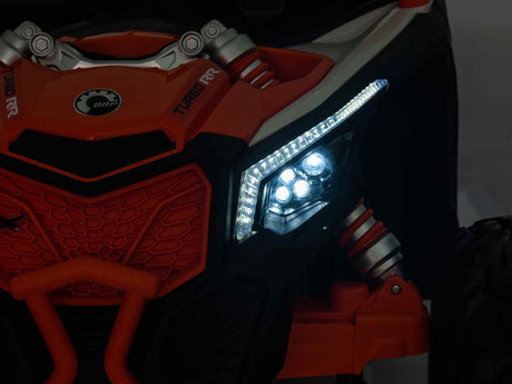 Close-up of the Black 2x24V/2WD Official Can-Am Maverick Ride on Buggy, LX Performance. The futuristic design showcases an illuminated LED headlight and decorative grill against a dimly lit background, highlighting the vehicles exciting front details.