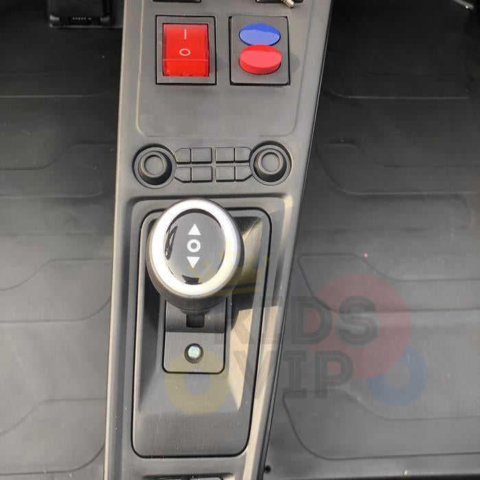 Close-up of the White 24V XXL SuperSport Big Kids Ride On Car control panel shows a circular gear shift with arrows, red and blue buttons above, and black circular buttons. A faint logo on the floor mat with colored circles enhances the realistic driving experience.