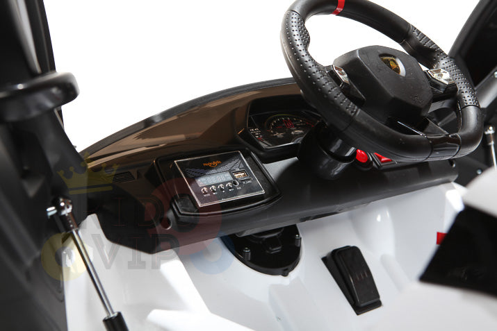 Close-up of a White 12V Lamborghini SVJ Sport Kids Ride-On Car dashboard featuring a black steering wheel, speedometer, and small digital display. The sleek interior has black and white accents, with visible pedals on the floor and part of a door on the left.