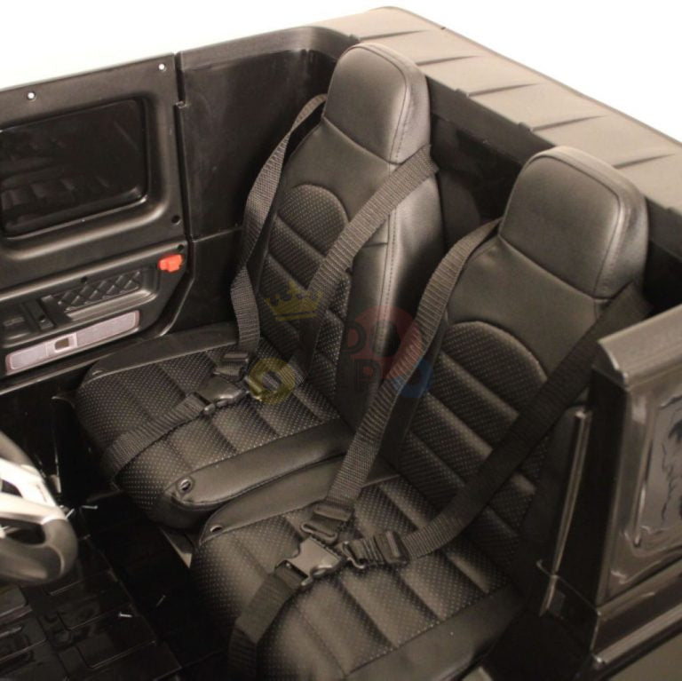 The Black 24V Mercedes Benz G63 Kids Ride On Car features two black plastic seats with detailed stitching and harness-style seat belts, a matte black cabin with detailed moldings, and is powered by a 24V battery system.