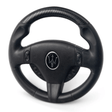 The Enhance Your Driving Experience with a 12v Maserati Gran Cabrio Steering Wheel features a black design, carbon fiber texture on top, a central trident logo, and two clusters of buttons, perfect for your Maserati.