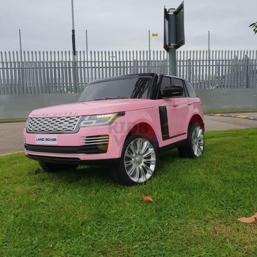 A Pink 24V Official Range Rover Complete MP4 Edition Kids Ride On Car with RC, featuring silver rims and a detailed grille, rests on lush green grass. A tall metal fence by a road with a signpost under an overcast sky forms the backdrop.