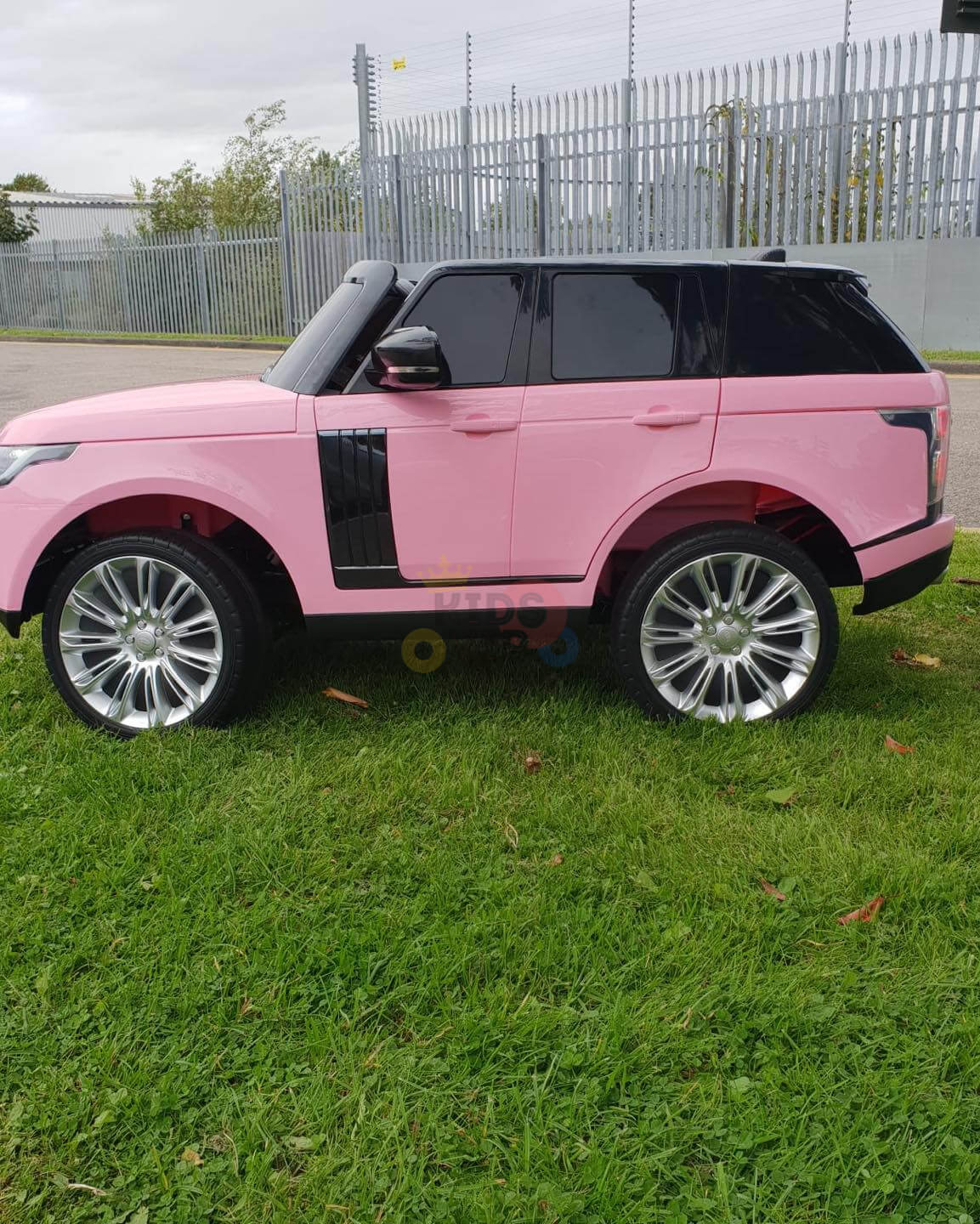 A Pink 24V Official Range Rover Complete MP4 Edition Kids Ride On Car with RC, styled like a mini-SUV, is parked on grass. It features shiny large wheels and dark tinted windows, with a metal fence and trees seen in the background.