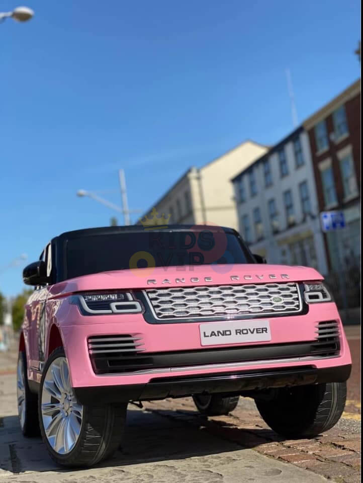 A Pink 24V Official Range Rover Complete MP4 Edition Kids Ride On Car with RC is parked on a cobblestone street, highlighting its detailed front grille and headlights. Range Rover and Land Rover logos are visible, set against buildings and a clear blue sky.