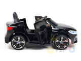 The Black Licensed BMW GT 12V Kids Ride On Power Car features realistic open doors, two seats, a steering wheel, and large wheels. It resembles a luxury vehicle and is positioned against a plain white background.