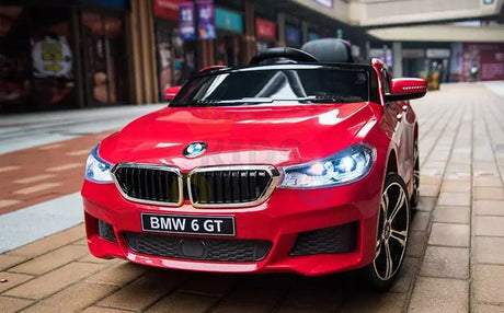 A Red 12V BMW GT Kids Licensed 1 Seater Ride On Car with Rubber Wheels and Remote is parked on a brick path. It features shining headlights and detailed design, resembling the real car, with blurred storefronts in the background enhancing the vibrant scene.