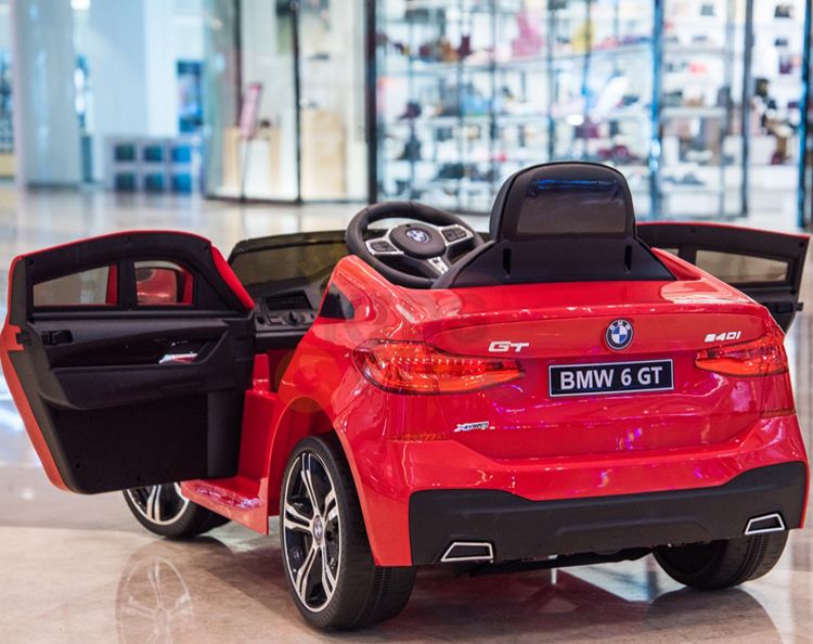 Displayed indoors on a shiny floor with open doors, the Red 12V BMW GT Kids Licensed 1 Seater Ride On Car features realistic details like a steering wheel and BMW 6 GT license plate. Store glass windows in the background enhance this licensed ride-ons appeal.
