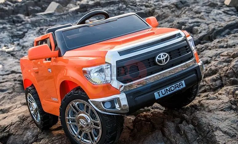 An Orange 12V Licensed Upgraded Toyota Tundra 1 Seater Kids Ride On Truck with RC is parked on rocky terrain, featuring realistic chrome accents, a front grille, and large tires, powered by a 12V battery for an enhanced off-road experience.