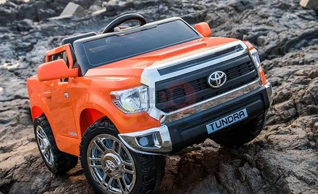 An Orange 12V Licensed Upgraded Toyota Tundra 1 Seater Kids Ride On Truck with RC is parked on rocky terrain, featuring realistic chrome accents, a front grille, and large tires, powered by a 12V battery for an enhanced off-road experience.