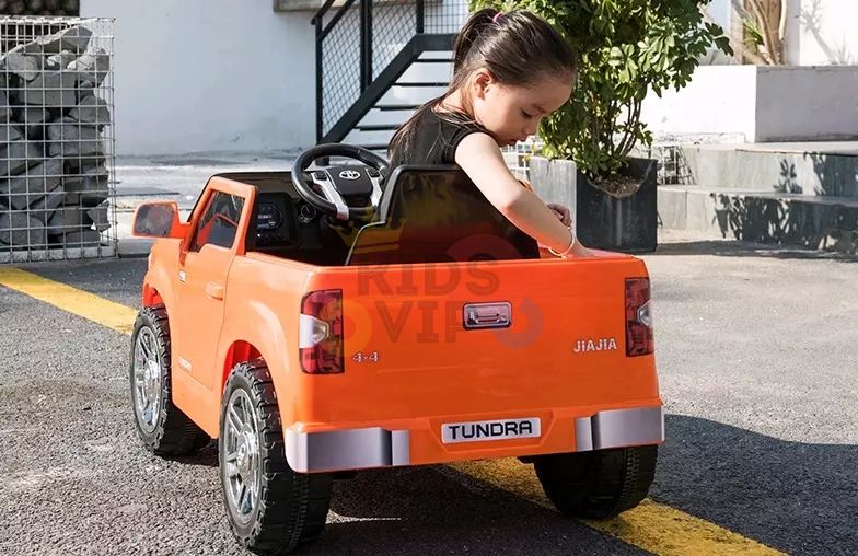 A child with a ponytail enjoys a sunny day in an Orange 12V Licensed Upgraded Toyota Tundra Kids Ride On Truck. Styled like a pickup, its parked on an outdoor surface with a yellow line, surrounded by plants and steps, creating a playful setting powered by its 12V battery.