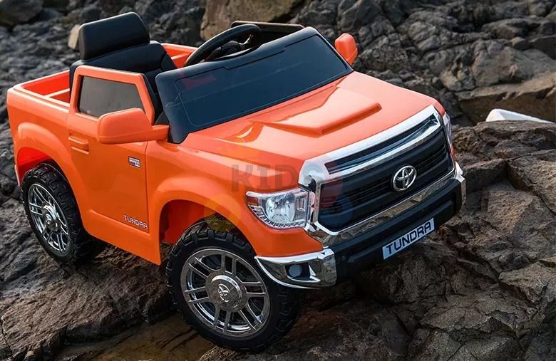 An Orange 12V Licensed Upgraded Toyota Tundra 1 Seater Kids Ride On Truck with RC, boasting detailed wheels, a black interior, and a rear cargo area, stands on rocky terrain. It features a TUNDRA badge and replicates the design and style of the real vehicle.