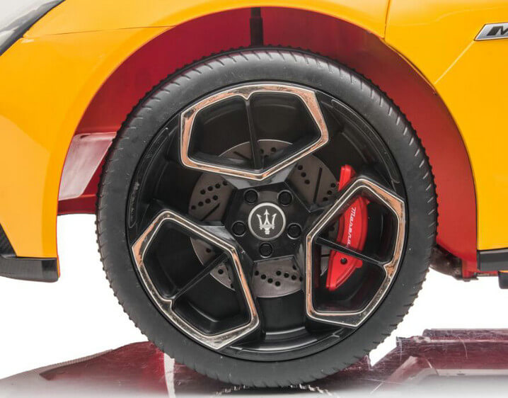 Close-up of Maserati MC20 wheel with black rim and angular silver outlines. Behind it, a red brake caliper hints at an exhilarating luxury ride. The cars body is glossy yellow. Product: Experience Luxury: Yellow XXL 24V/105W Maserati MC20 | Premium 2-Seater with RC, Leather Seats & Rubber Tires.