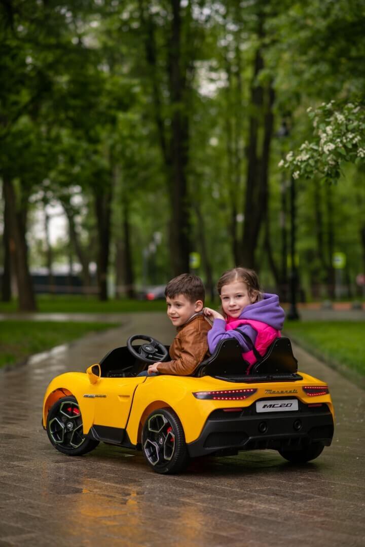 Two children cruise through a park in a yellow ride-on vehicle on a wet path, surrounded by lush green trees. The boy steers in his brown jacket while the girl sits beside him in pink and purple, enjoying their mini adventure in an Experience Luxury: Pink XXL 24V/105W Maserati MC20.