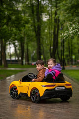 Two kids relish a luxury ride in the Experience Luxury: Yellow XXL 24V/105W Maserati MC20, cruising on a rainy park path lined with tall green trees. The boy in brown takes the wheel, and the girl in pink and purple sits beside him, enjoying their premium 2-seater with leather seats and rubber tires.