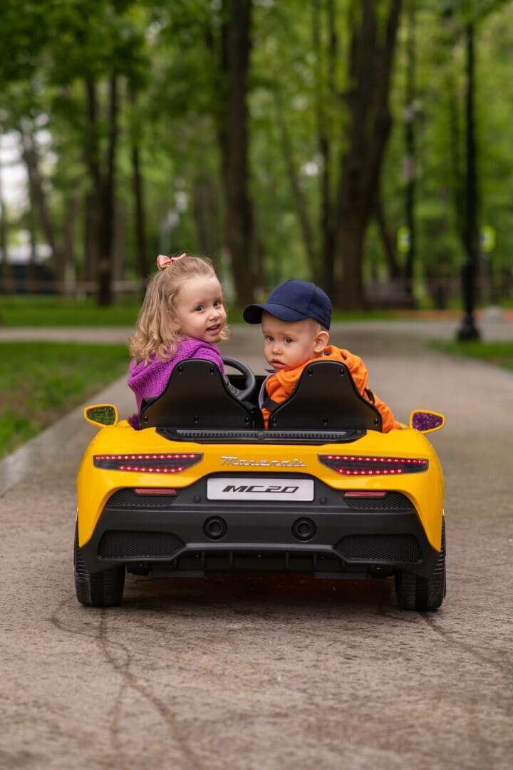 Two kids ride a small yellow vehicle in the park; one wears a purple jacket, the other an orange jacket and blue cap. They look back at the camera, surrounded by green trees, capturing the charm of a Pink XXL 24V Maserati MC20 luxury mini car adventure with leather seats and rubber tires.