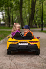 Two young children enjoy a ride in the Experience Luxury: Yellow XXL 24V /105W Maserati MC20, a premium 2-seater with RC, leather seats, and rubber tires. The girl in her purple jacket smiles at the boy beside her, his orange jacket and cap standing out against the parks lush backdrop.