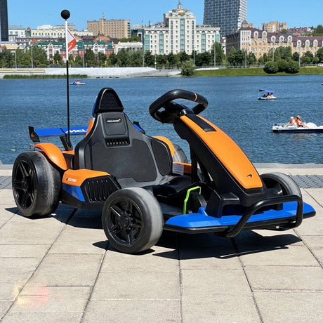 The Orange 24V Official Complete Edition McLaren Super Drifting Go-Kart is stationed on paved ground by a peaceful river, with buildings, trees, and paddle boats. Its vibrant colors add excitement to the clear, sunny setting—ideal for a thrilling go-kart drifting adventure.