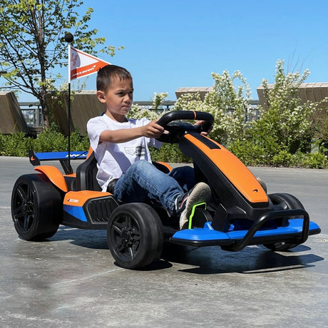 A boy in a white shirt and jeans enjoys an exhilarating ride on the Orange 24V Official Complete Edition McLaren Super Drifting Go-Kart, featuring a small flag, with lush plants and modern buildings beneath the vast blue sky.