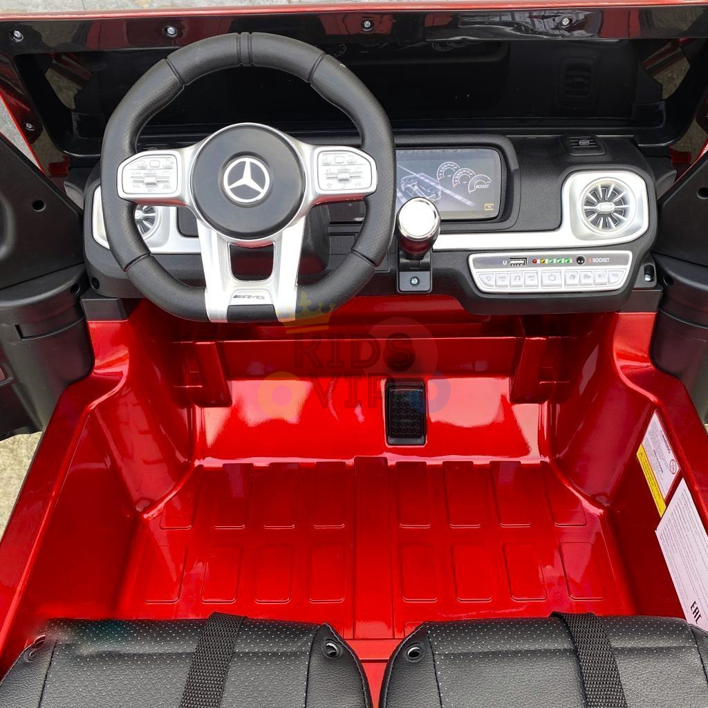 The 2 Seats 4x4 Official Mercedes Benz G63 Complete Edition Red 24V Kids Ride On Car with RC mimics a real G63, featuring a dashboard with speedometer, control buttons, and steering wheel. It has black seats on a red floor and runs on a powerful 24V battery for an authentic driving experience.