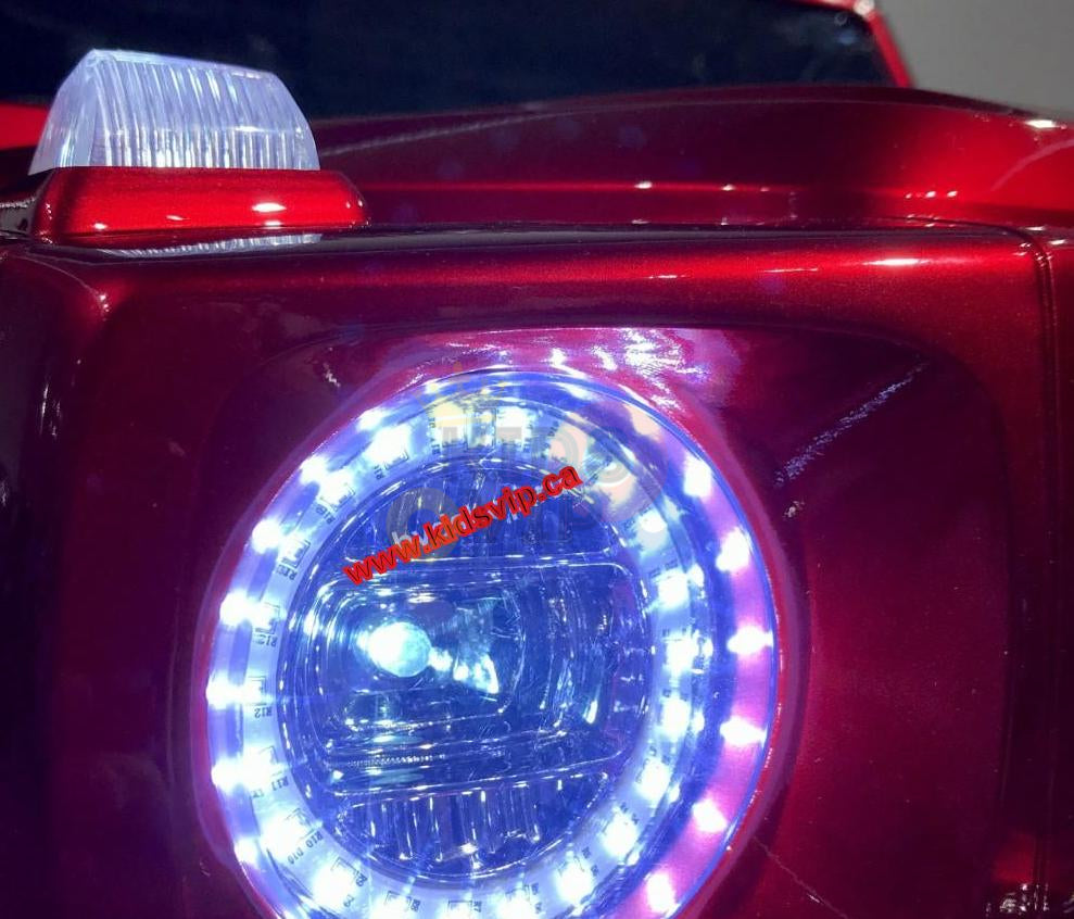 A close-up of the red 2 Seats 4x4 Official Mercedes Benz G63 Complete Edition Kids Ride On Car shows its circular headlight with bright LEDs and a reflective side marker above. A website URL subtly appears at the image center, accentuating this toy cars sleek design.