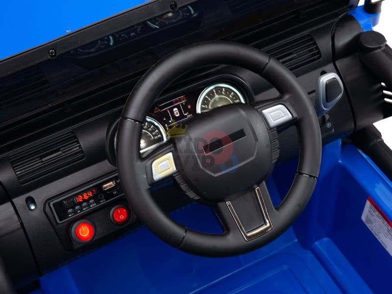 A close-up of the interior of the Blue 12V Sport Edition Trailcat Kids Ride-on Truck shows a black steering wheel, dashboard with circular dials, and a mix of red and black buttons on its blue panel. Ideal for an adventurous experience!.